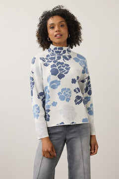 A woman with curly dark hair wearing a white mock-neck long-sleeve top with a blue floral pattern, paired with gray jeans, standing against a light gray studio background.