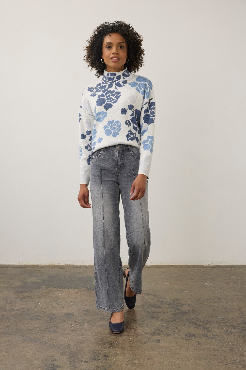 A woman modeling a white mock-neck knit sweater with a blue floral pattern, paired with wide-leg gray jeans and navy flats, standing against a plain off-white wall.