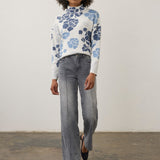 A woman modeling a white mock-neck knit sweater with a blue floral pattern, paired with wide-leg gray jeans and navy flats, standing against a plain off-white wall.