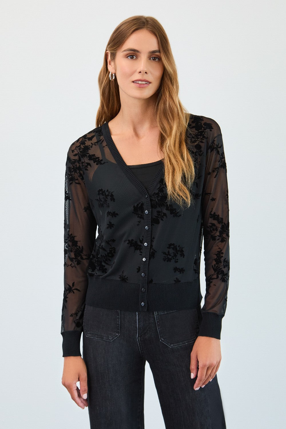 Woman wearing a black floral lace cardigan over a black top with blue jeans on a white background