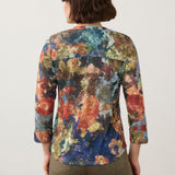 Woman wearing a multi coloured 3/4 sleeve henley top with green jeans on a light background.