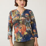 Woman wearing a multi coloured 3/4 sleeve henley top with green jeans on a light background.