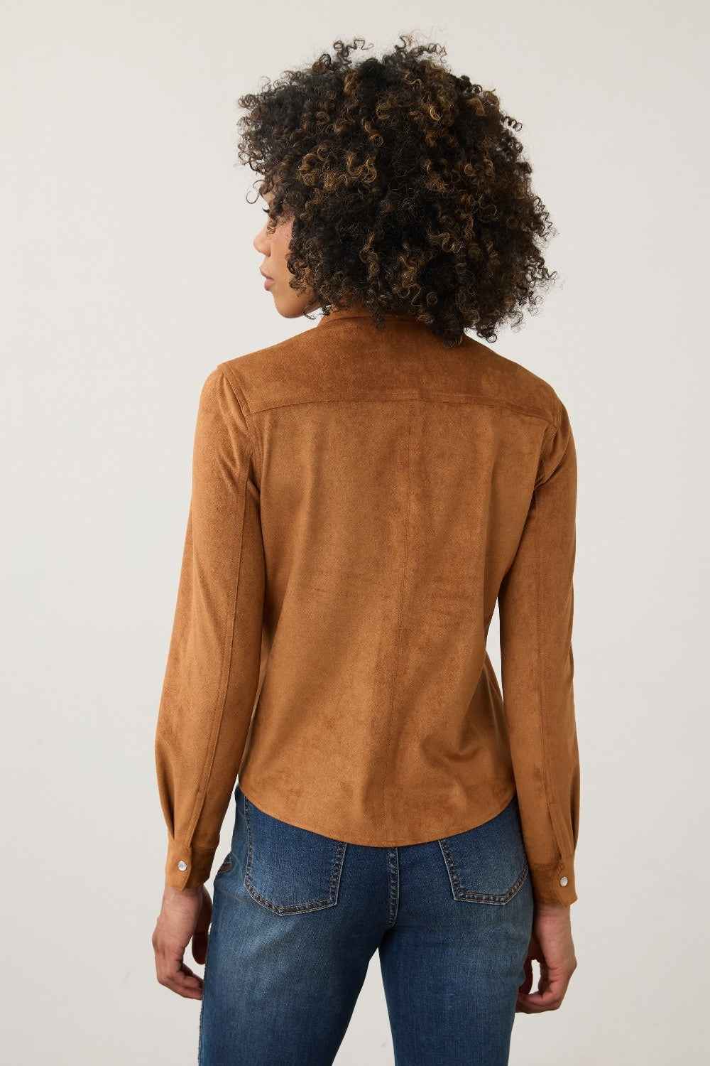 A woman with curly dark hair is shown from the back wearing a tan suede long-sleeve shirt and blue jeans against a plain light gray studio background.