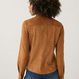 A woman with curly dark hair is shown from the back wearing a tan suede long-sleeve shirt and blue jeans against a plain light gray studio background.