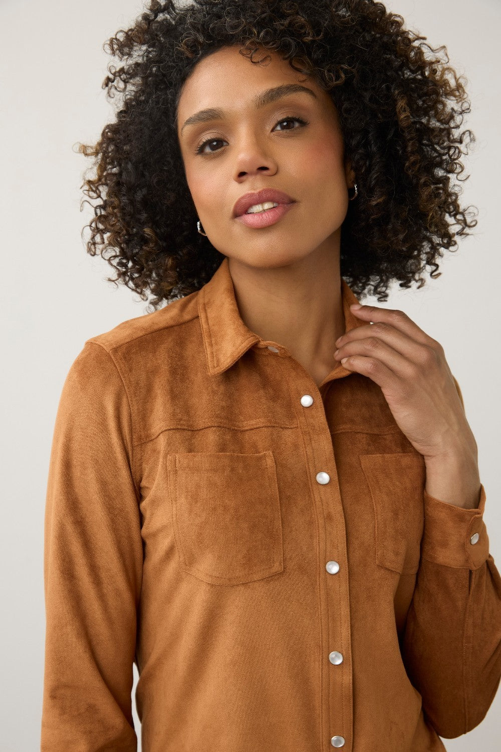 A woman with curly dark hair wearing a brown suede long-sleeve button-up shirt with two chest pockets, photographed against a light gray studio background.