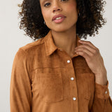 A woman with curly dark hair wearing a brown suede long-sleeve button-up shirt with two chest pockets, photographed against a light gray studio background.
