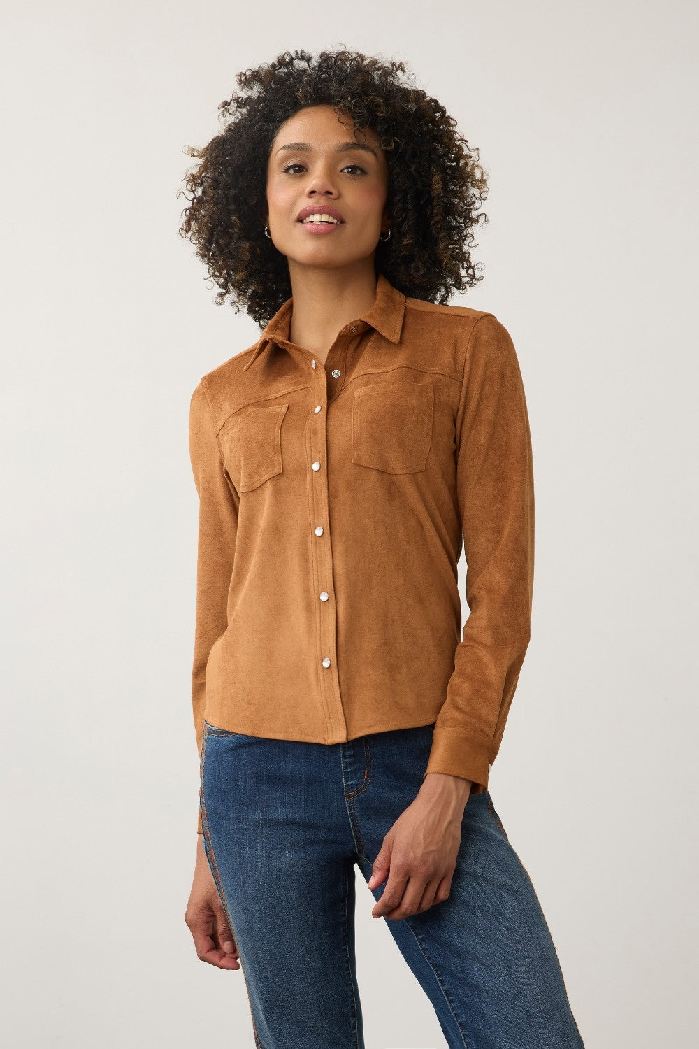A woman with curly dark hair wearing a tan suede long-sleeve button-up shirt and blue jeans, standing against a light gray studio background.