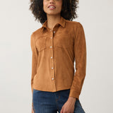 A woman with curly dark hair wearing a tan suede long-sleeve button-up shirt and blue jeans, standing against a light gray studio background.
