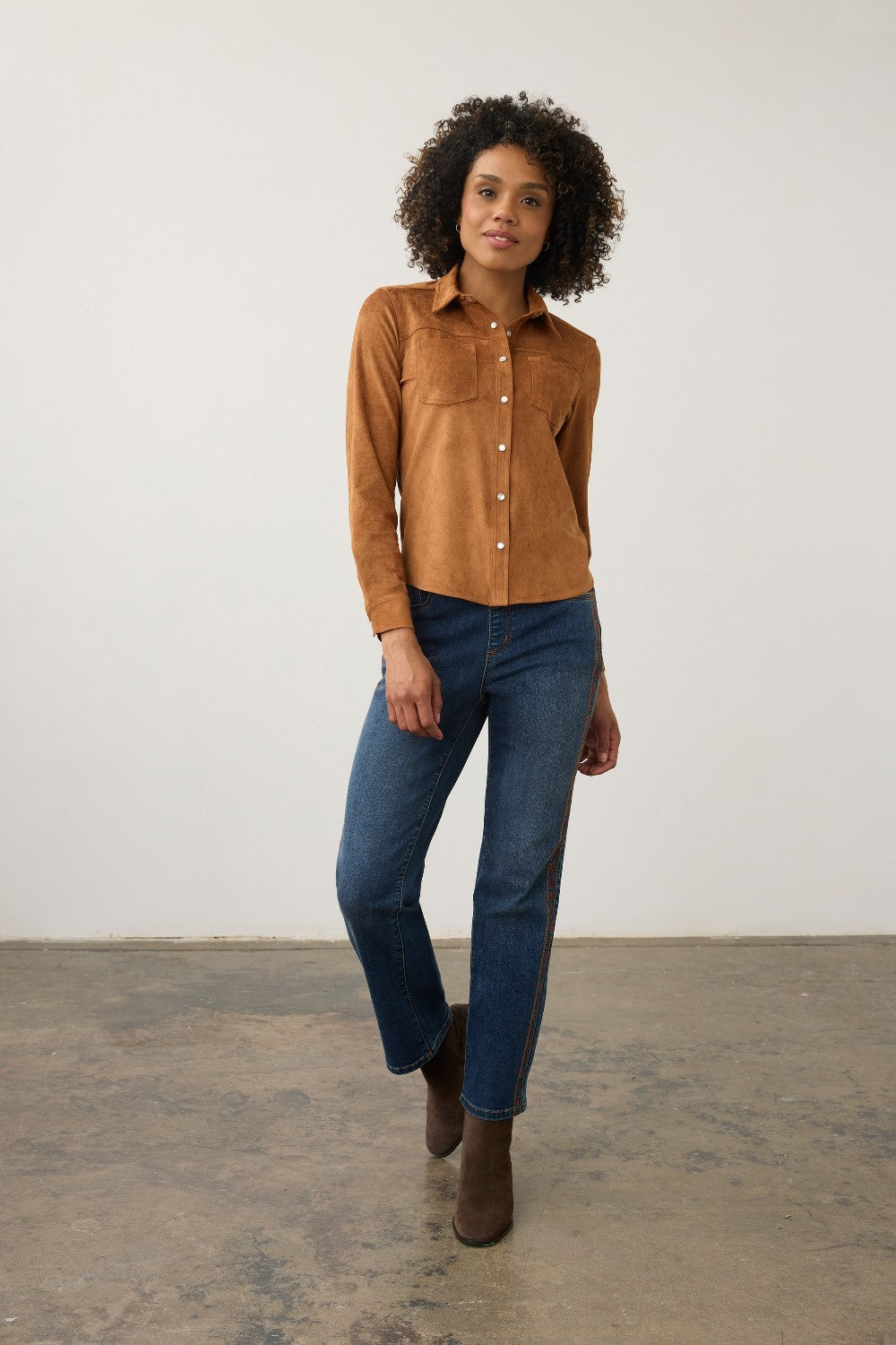 A woman modeling a tan suede long-sleeve button-up shirt with blue jeans and brown ankle boots, standing against a plain off-white wall and concrete floor.