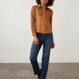 A woman modeling a tan suede long-sleeve button-up shirt with blue jeans and brown ankle boots, standing against a plain off-white wall and concrete floor.