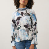 A woman with curly dark hair wearing a long-sleeve blouse with a blue, white, and black floral watercolor print, paired with blue jeans, standing against a light gray studio background.