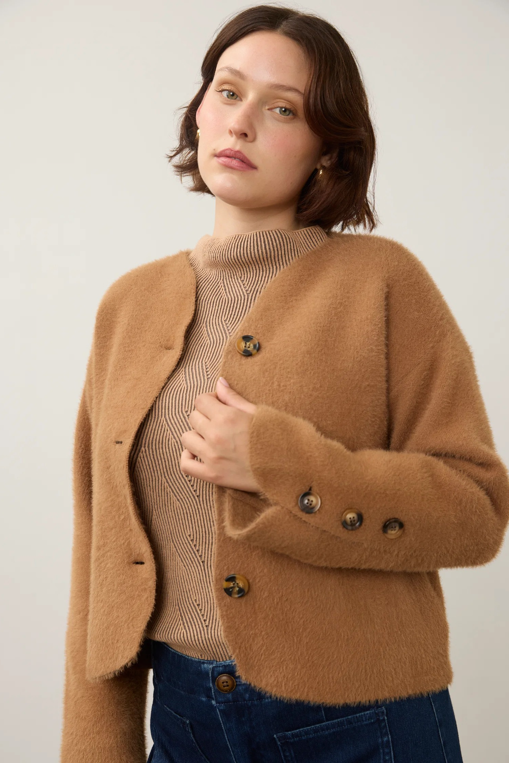 A woman wearing a fuzzy camel brown cropped cardigan over a beige ribbed turtleneck sweater and dark blue jeans, photographed against a plain light background.