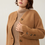 A woman wearing a fuzzy camel brown cropped cardigan over a beige ribbed turtleneck sweater and dark blue jeans, photographed against a plain light background.