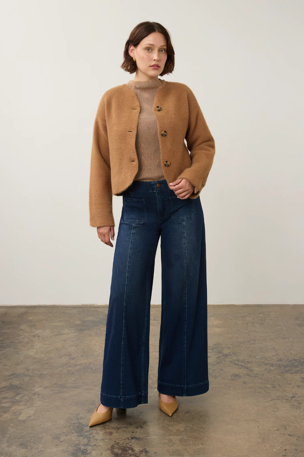 A model stands in a studio wearing a camel cropped button-front cardigan over a beige ribbed mock-neck top, paired with dark blue wide-leg jeans and tan pointed-toe heels.