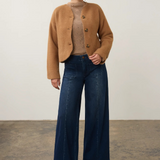 A model stands in a studio wearing a camel cropped button-front cardigan over a beige ribbed mock-neck top, paired with dark blue wide-leg jeans and tan pointed-toe heels.