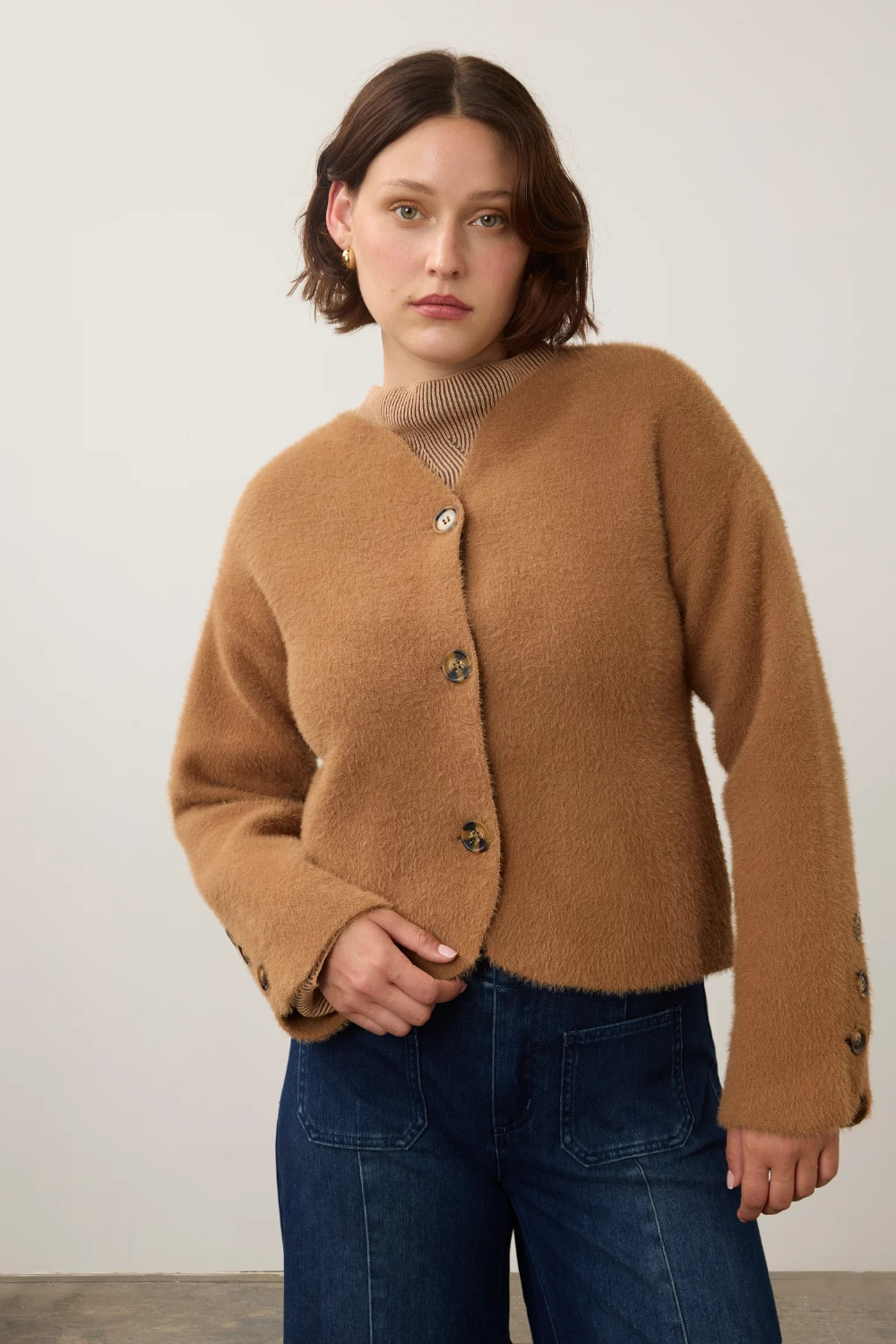 A woman wearing a cropped, fuzzy camel-brown cardigan with decorative buttons, paired with dark blue jeans, standing against a plain off-white wall.