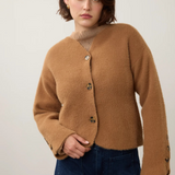 A woman wearing a cropped, fuzzy camel-brown cardigan with decorative buttons, paired with dark blue jeans, standing against a plain off-white wall.