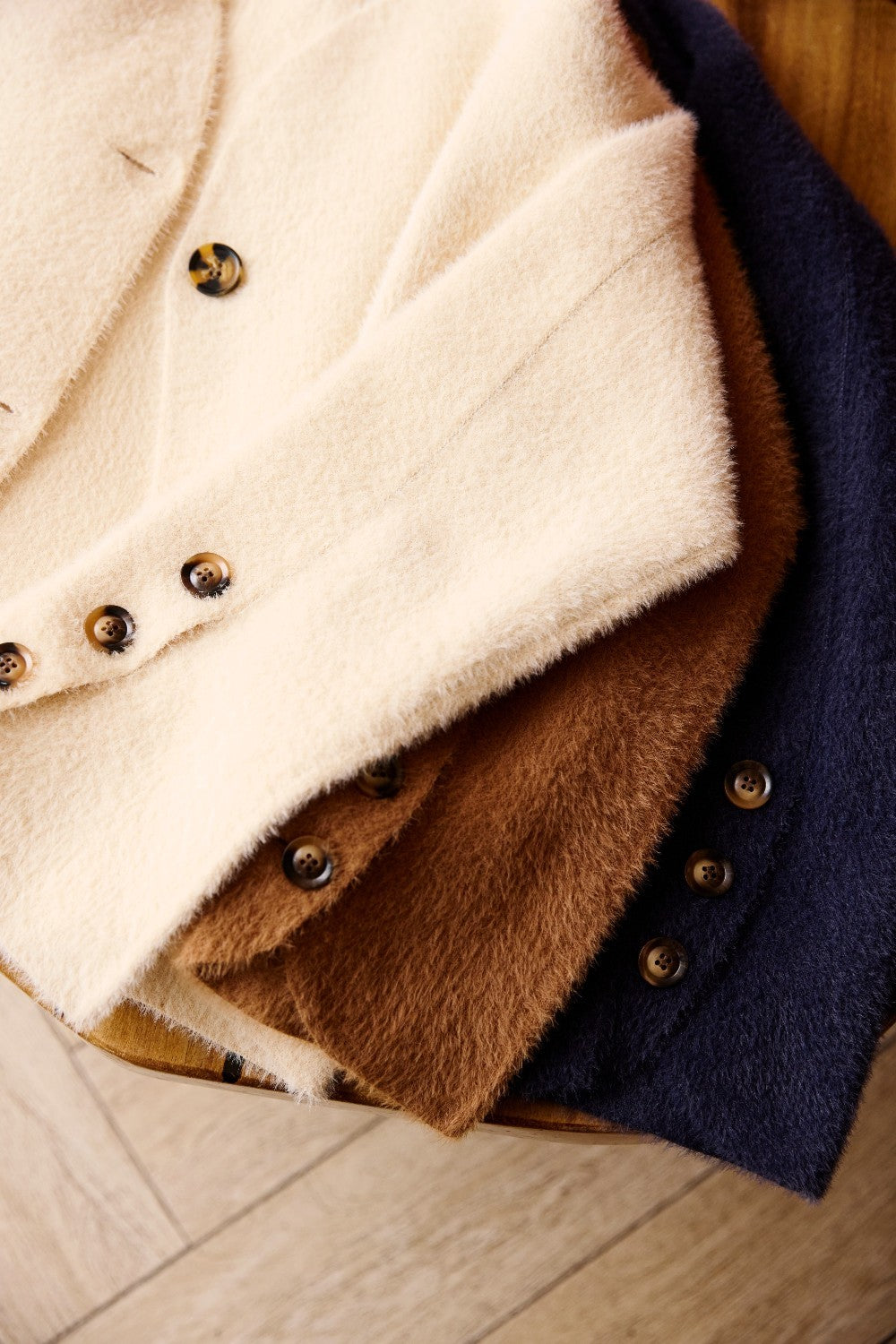 Three soft fuzzy button-front jackets stacked on a wooden surface, shown in cream, brown, and navy.