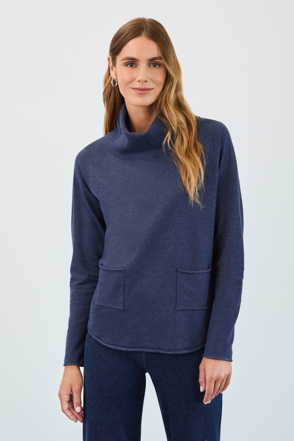 Woman wearing a navy blue cowl-neck sweater with pockets on a light gray background