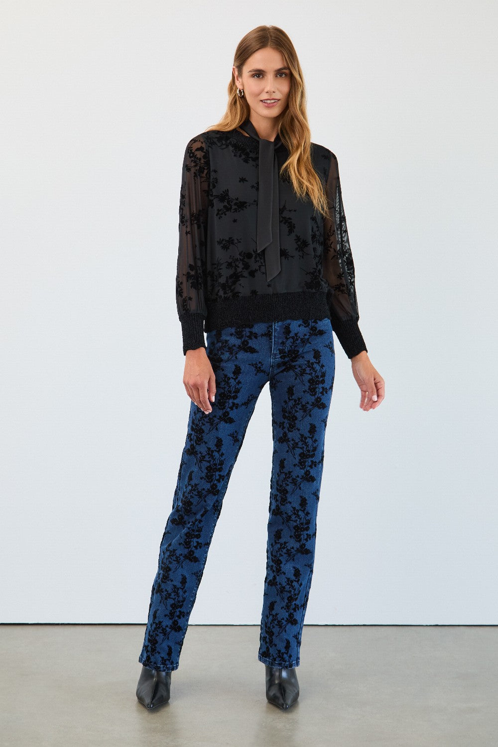 Woman wearing a black floral blouse and blue pants against a white background
