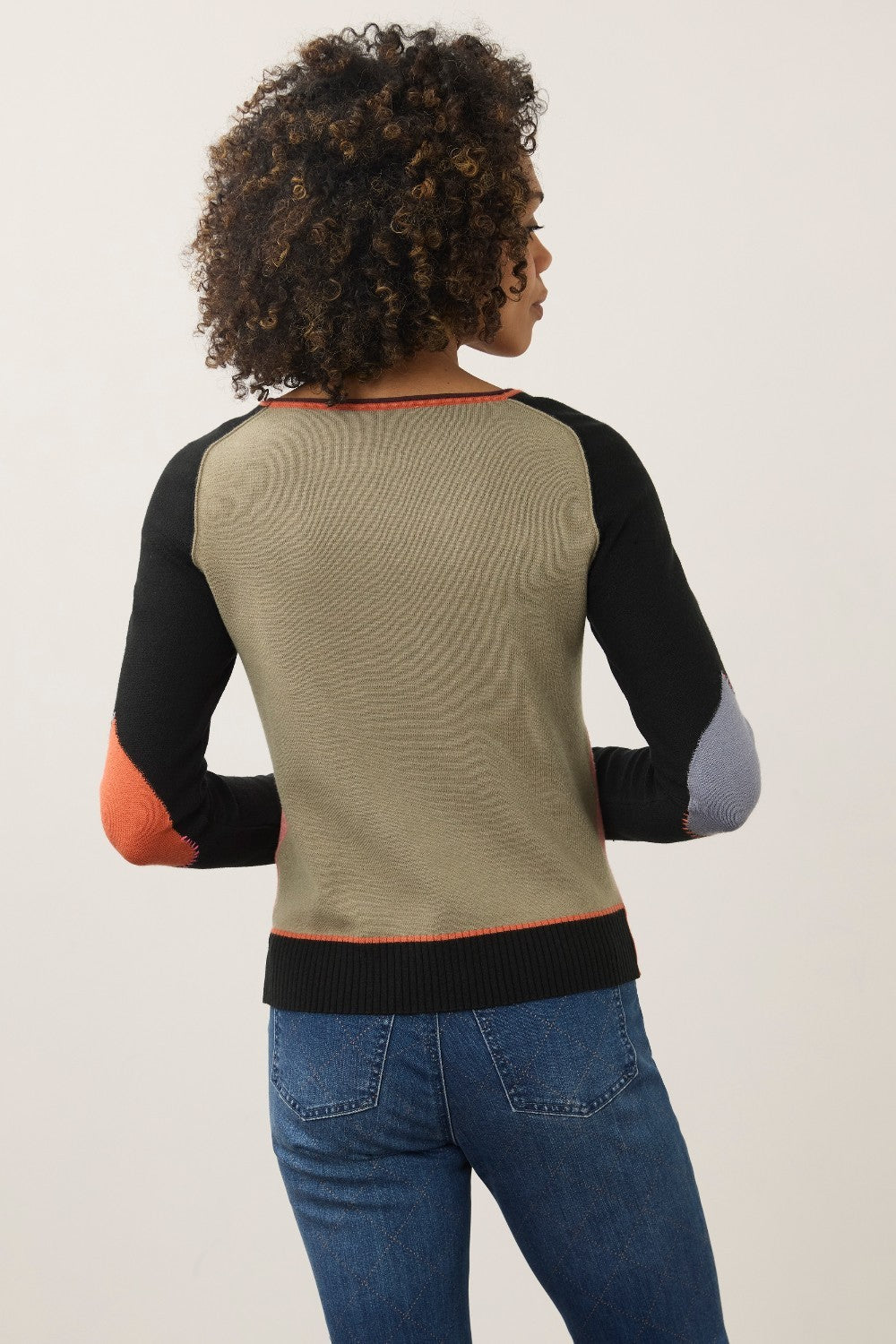 A woman shown from the back wearing a color-blocked knit sweater with a beige back panel, black raglan sleeves, orange elbow patches, and a gray elbow patch, paired with blue jeans against a plain light background.