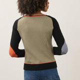 A woman shown from the back wearing a color-blocked knit sweater with a beige back panel, black raglan sleeves, orange elbow patches, and a gray elbow patch, paired with blue jeans against a plain light background.