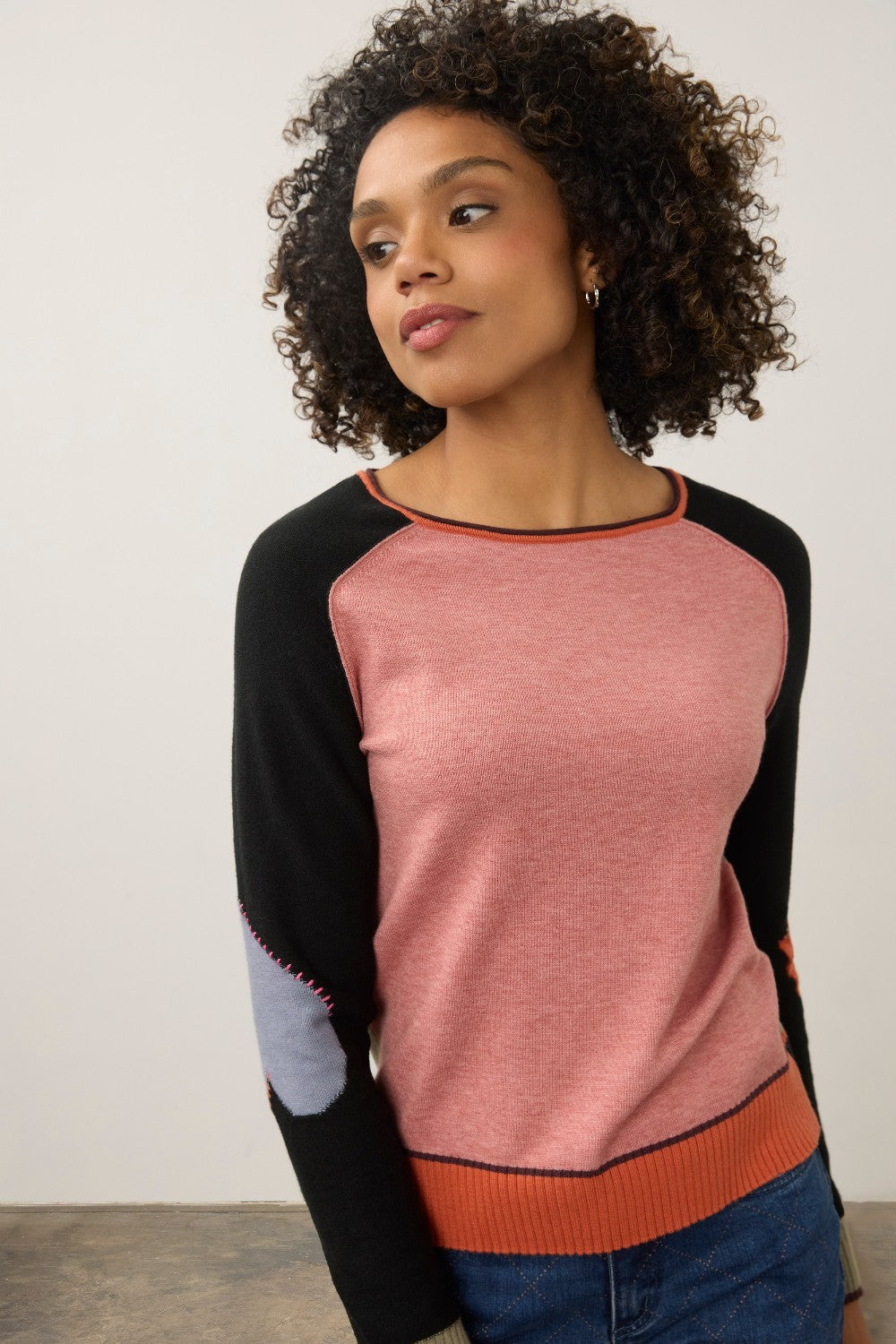 A woman wearing a color-blocked knit sweater with a heathered pink body, black raglan sleeves, and orange ribbed trim, paired with blue jeans against a plain light gray wall.