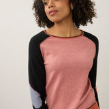 A woman wearing a color-blocked knit sweater with a heathered pink body, black raglan sleeves, and orange ribbed trim, paired with blue jeans against a plain light gray wall.