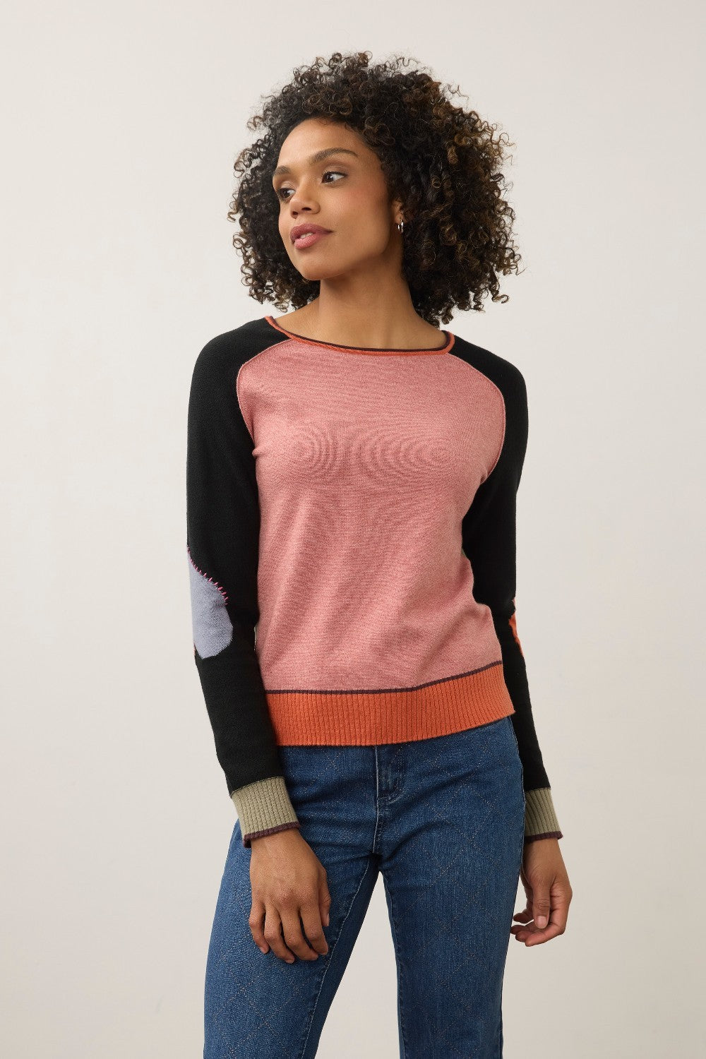 A woman modeling a color-blocked long-sleeve knit sweater with a dusty pink body, black raglan sleeves, and an orange ribbed hem and underarm band, paired with blue jeans against a plain light gray studio background.