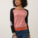 A woman modeling a color-blocked long-sleeve knit sweater with a dusty pink body, black raglan sleeves, and an orange ribbed hem and underarm band, paired with blue jeans against a plain light gray studio background.