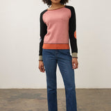 A woman modeling a colorblock knit sweater with a pink body, black sleeves, and orange ribbed trim, paired with blue jeans and black loafers, standing against a plain studio wall.