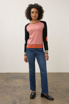 A woman modeling a colorblock knit sweater with a pink body, black sleeves, and orange ribbed trim, paired with blue jeans and black loafers, standing against a plain studio wall.