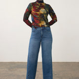 Woman wearing a printed mock neck top with blue jeans and brown shoes on a neutral background.