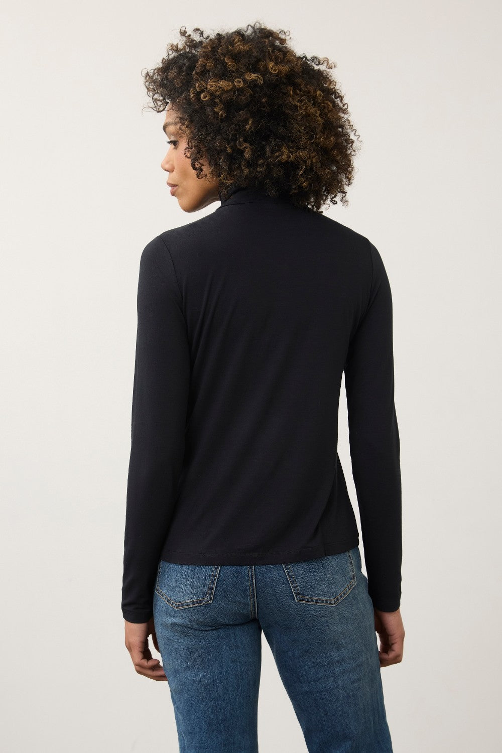 A woman with curly brown hair is shown from the back wearing a black long-sleeve mock neck top and blue jeans against a plain light gray studio background.