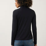 A woman with curly brown hair is shown from the back wearing a black long-sleeve mock neck top and blue jeans against a plain light gray studio background.