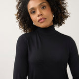 A woman with curly dark hair wearing a black long-sleeve mock neck turtleneck top, photographed against a plain light gray studio background.