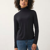 A woman with curly dark hair wearing a black long-sleeve mock neck turtleneck top and blue jeans, standing against a plain light gray studio background.