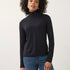 A woman with curly dark hair wearing a black long-sleeve mock neck turtleneck top and blue jeans, standing against a plain light gray studio background.