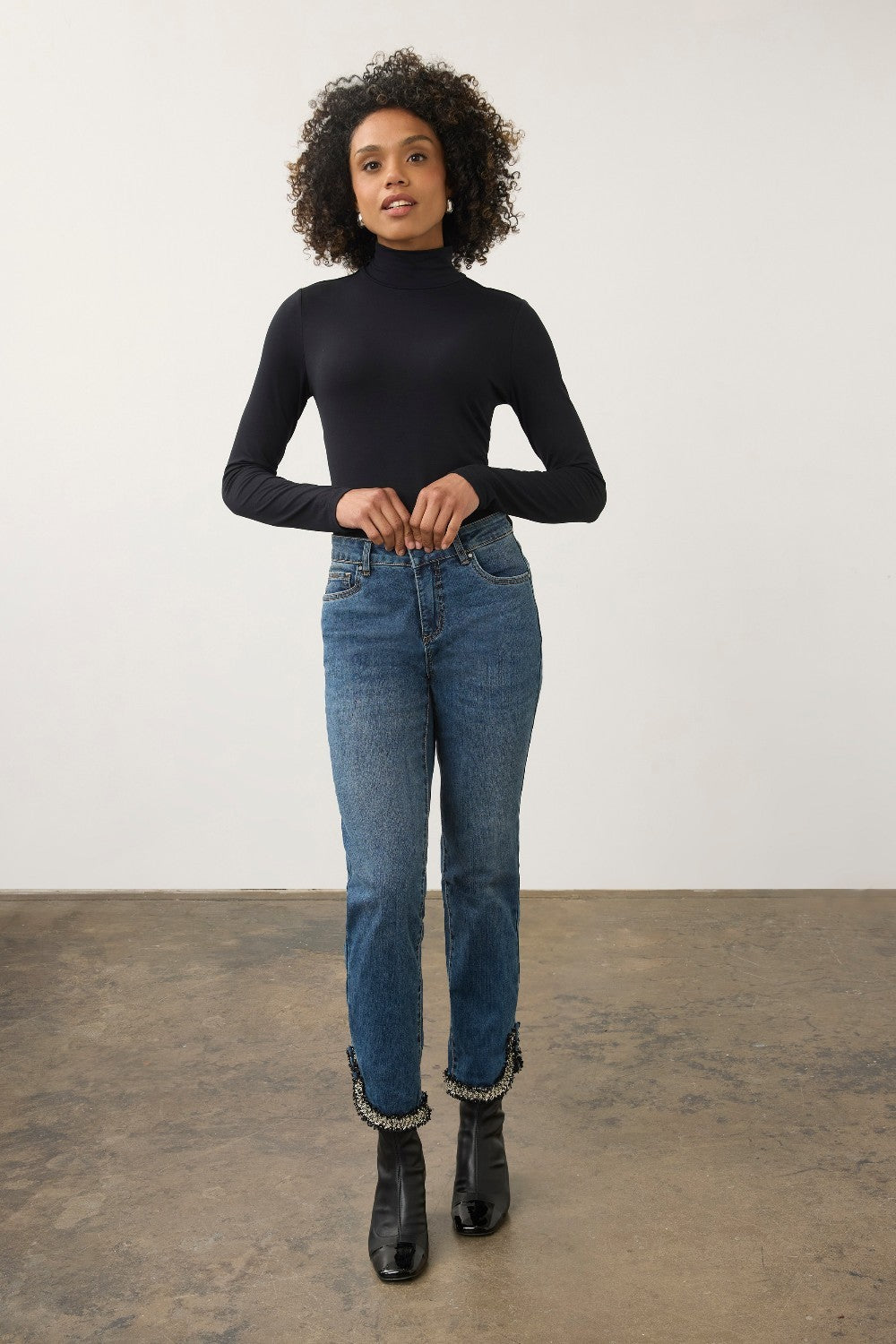 A woman stands in a studio wearing a fitted black long-sleeve turtleneck top, blue jeans with frayed hems, and black ankle boots against a plain light wall and concrete floor.