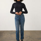 A woman stands in a studio wearing a fitted black long-sleeve turtleneck top, blue jeans with frayed hems, and black ankle boots against a plain light wall and concrete floor.