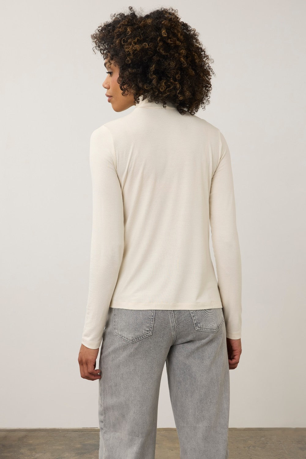 A woman with curly dark hair wearing a cream-colored long-sleeve mock neck top and light gray jeans, standing against a plain light gray studio background.