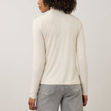 A woman with curly dark hair wearing a cream-colored long-sleeve mock neck top and light gray jeans, standing against a plain light gray studio background.