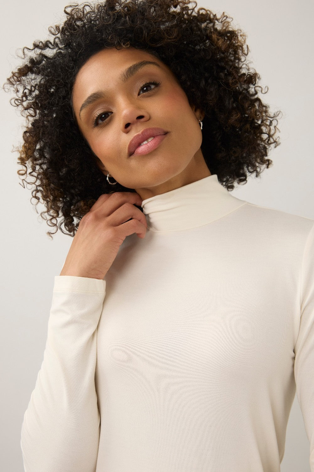 A woman with curly dark hair wearing a cream-colored long-sleeve mock neck top against a plain light gray studio background.