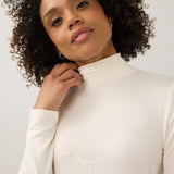 A woman with curly dark hair wearing a cream-colored long-sleeve mock neck top against a plain light gray studio background.