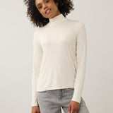 A woman with curly dark hair wearing a cream-colored long-sleeve mock neck top and light gray jeans, standing against a plain light gray studio background.