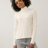 A woman with curly dark hair wearing a cream-colored long-sleeve mock neck top and light gray jeans, standing against a plain light gray studio background.