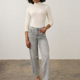 A woman with curly dark hair wearing a cream-colored long-sleeve mock neck top, light gray jeans and cream loafers, standing against a plain light gray studio background.