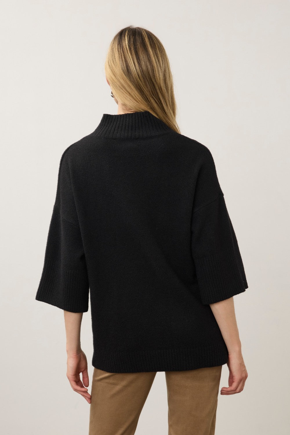 A woman viewed from the back wearing a black knit mock-neck sweater with three-quarter sleeves and tan pants, set against a plain light gray studio background.