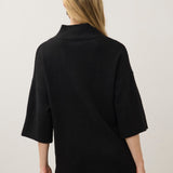 A woman viewed from the back wearing a black knit mock-neck sweater with three-quarter sleeves and tan pants, set against a plain light gray studio background.