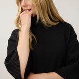 A woman with blonde hair wearing a black ribbed mock-neck sweater, posing with her hand near her chin against a plain light gray background.